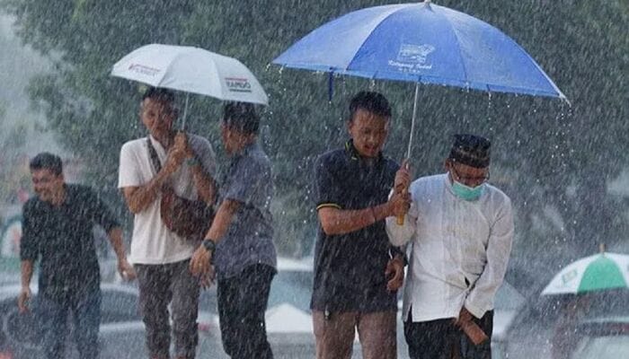 BMKG Prediksi Hujan Ringan, Sekelompok orang berjalan di tengah hujan lebat sambil menggunakan payung di jalanan kota.