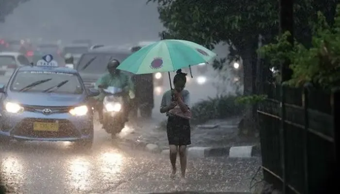 Mayoritas Wilayah Indonesia Diprediksi Hujan, Seorang wanita berjalan kaki di tengah hujan deras dengan payung hijau, di jalanan kota yang dipenuhi kendaraan bermotor.
