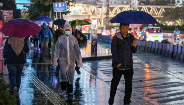 Cuaca Awal Agustus, Orang-orang berjalan menggunakan payung dan jas hujan di trotoar kota saat hujan turun di malam hari.