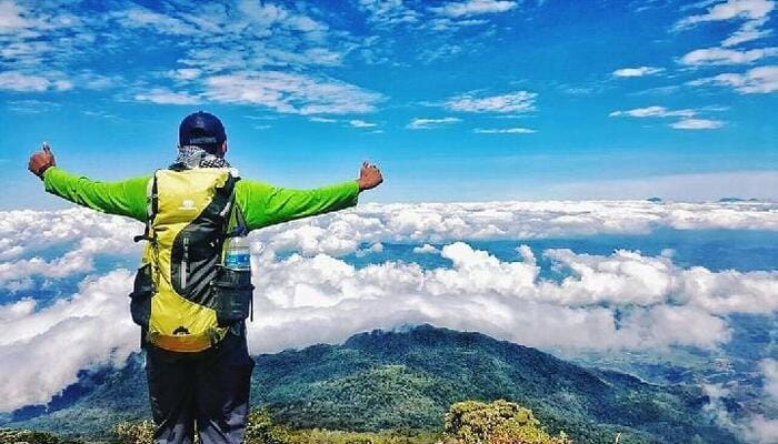 Gunung Lewotobi Erupsi, Seorang pendaki dengan jaket hijau dan ransel berdiri di puncak Gunung Ciremai, membentangkan tangan sambil menikmati pemandangan awan putih dan langit biru.