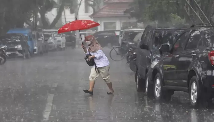 Cuaca Senin Mencekam, Seorang perempuan menyeberang jalan dengan payung merah saat hujan deras di tengah deretan mobil terparkir.